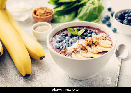 Acai smoothie Schüssel auf grauem Beton Hintergrund. Smoothie mit superfood toppings Banane, Nussbaum, Kokos, Chia Samen und frische Blaubeeren. Selektive Stockfoto