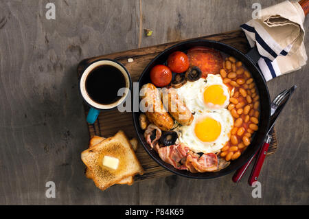 Englisches Frühstück mit Würstchen, Speck, Rührei, Bohnen, Toast und Kaffee auf Holztisch, Ansicht von oben. Im rustikalen Stil Stockfoto