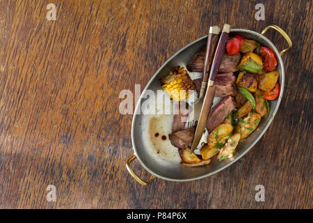 Roastbeef Steak mit gebratenem Gemüse (Kartoffeln, Mais, Paprika, Tomaten, Kürbis) in Kupfer auf einem Holztisch mit Rand, Ansicht von oben. Herbstliche Stimmung Stockfoto