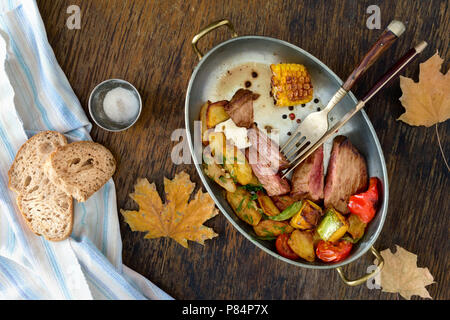 Roastbeef Steak mit gebratenem Gemüse (Kartoffeln, Mais, Paprika, Tomaten, Kürbis) in der Pfanne auf einem Holztisch. Herbstliche Stimmung. Ansicht von oben Stockfoto