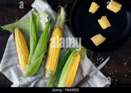 Frische Maiskolben auf dunklen Holztisch mit Grillpfanne, Ansicht von oben Stockfoto