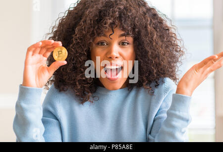 African American woman holding golden Bitcoin cryptocurrency zu Hause sehr glücklich und aufgeregt, Sieger Ausdruck feiern Sieg schreien mit großen Stockfoto