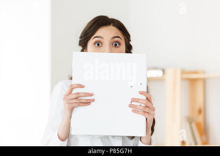 Junge Frau mit überraschten Augen spähen hinter Papier Poster. Geschäftsfrau, die große weiße Banner in modernen Büro. Stockfoto