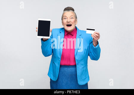 Gealterte Frau zeigen tablet Bildschirm und Kreditkarte und Suchen von der Kamera begeistert. Großmutter im blauen Anzug mit gesammelten graue Haare bun Frisur. Ich Stockfoto