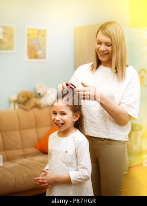 Mutter ist das Flechten der Haare stehen ihre junge Tochter im Zimmer der Kinder. Glücklich liebende Familie. Stockfoto