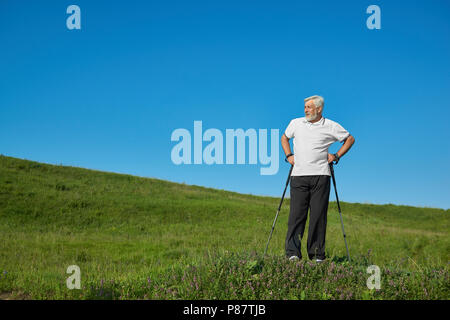 Mann, der sportsuit stehend mit Tracking klebt auf dem Grünen Hügel. Tragen weiße Poloshirt mit dunkelblauen Streifen, schwarze Hose, Turnschuhe. Auf der Suche nach einem Stockfoto
