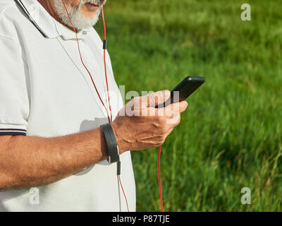 Foto von alter Mann Kontrolle Zeit beschnitten, während draußen laufen. Halten Handy, auf sportstyle handwatch. Tragen klassisches weißes Poloshirt Esprit Stockfoto