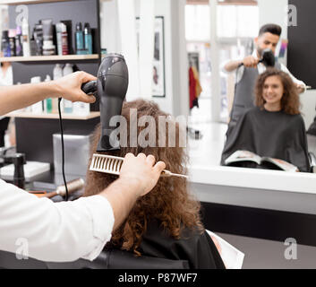 Rückansicht Blickrichtung von Curly hairstyler Trocknen weibliche client Haare. Unscharfe Reflexion in den großen Spiegel. Mit Hilfe von Pinsel und Fön. Tragen weiße Hemd, g Stockfoto