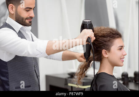 Seitenansicht des Arabischen hairstyler Trocknen lächelnd lockiges Haar der weiblichen Kunden. Unscharfe Reflexion in den großen Spiegel. Mit Hilfe von Pinsel und Fön. Wearin Stockfoto