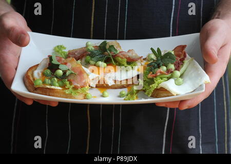 Bruschetta mit Rucola, chrispy Speck und pochiertem Ei auf einem weißen Teller serviert Stockfoto