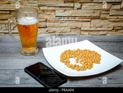 Auf einer hölzernen Tisch ist ein großes Glas Bier, eine Platte von Muttern und ein Telefon. Stockfoto