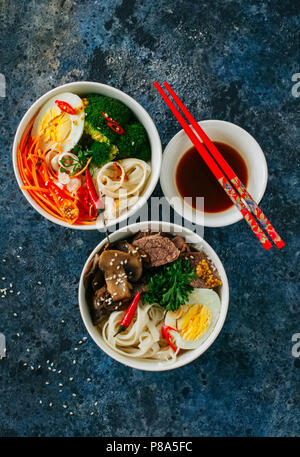 Asiatische Lebensmittel Konzept. Zwei verschiedene Schüsseln mit Udon-nudeln. Mit Brokkoli, Ei, Karotte und Gewürzen. Andere mit Fleisch, Pilzen, Ei und Petersilie. T Stockfoto