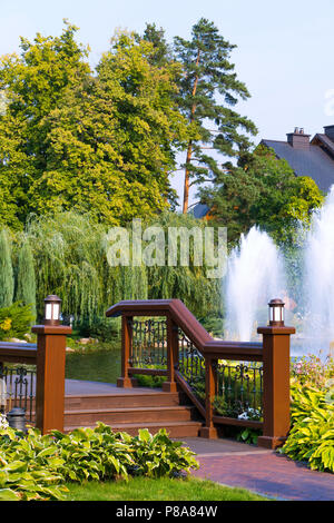 Eine moderne Holzbrücke über den See mit Springbrunnen, von hohen grünen Bäumen mit einem Cottage und einem blauen, wolkenlosen Himmel im Hintergrund umgeben. Für Stockfoto