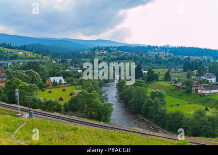 Dorf auf einem grünen Ebene mit einem Bahnhof und einem Berg River vor dem Hintergrund der fernen Gipfeln und einem grauen bewölkten Himmel. Ort der Erholung und des Tourismus. Fo Stockfoto