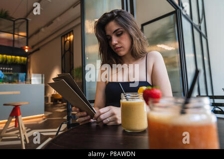 Reizende junge Dame in casual Outfit Menü Auslesen beim Sitzen im Cafe Tisch in der Nähe Gläser smoothie Stockfoto