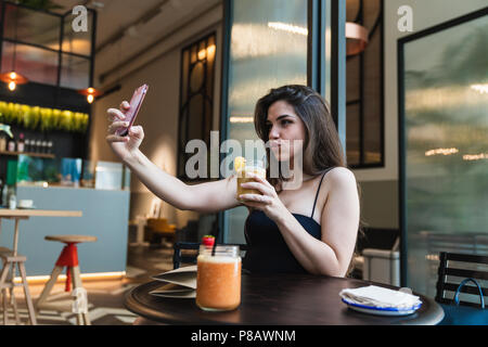 Reizende junge Dame im stilvollen Outfit holding Glas frischen Smoothie und posieren für selfie während am Tisch in schönen Cafés sitzen Stockfoto