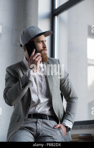 Bärtige Geschäftsmann in Baseball Cap sprechen von Smartphone und Wegsehen Stockfoto