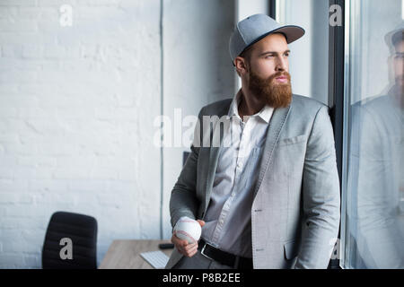 Bärtige Geschäftsmann im Baseball Kappe am Fenster schauen und Holding baseball ball Stockfoto