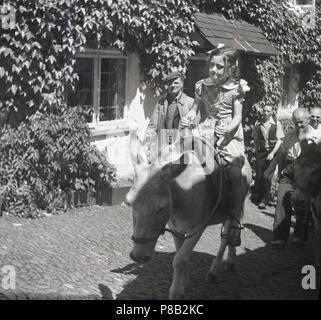 1950er Jahre, ein junges Mädchen auf einem Esel in Clovelly, Devon, England, UK. Jahrhundert Fischerdorf hat schmalen, steilen Straßen und so für Jahrhunderte Esel wurden für Waren und Leute aufstehen und auf der Straße verwendet. Stockfoto