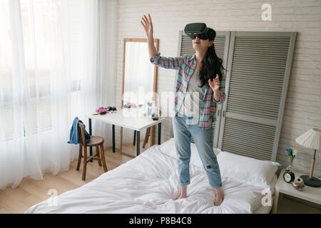 Asiatische Frau spielen der VR-Gerät auf ihrem Bett, die Arme, die sich in der Luft, als wie sie ist ergreifend und fangen etwas der realen Welt. Stockfoto