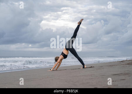 Sportliche junge Frau Yoga in einem vierbeinigen Downward-Facing Hund darstellen (Eka Pada Adho Mukha Svanasana) an der Küste Stockfoto