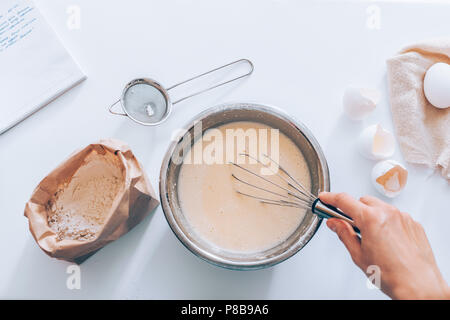 Prozess der Herstellung der Teig, der Frau Hand peitschen Eier und Mehl in Schüssel neben dem Handgeschriebenen Rezept- und Sieb, Ansicht von oben. Flach Zusammensetzung der ing Stockfoto