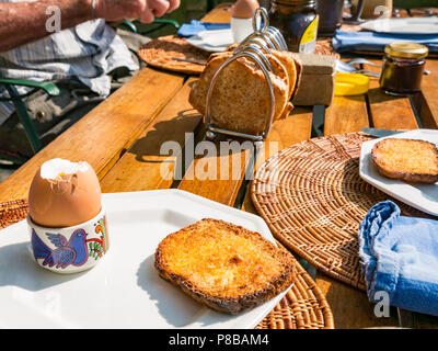 Mann essen Frühstück im Freien während der Sommerhitze, mit gekochtem Ei und Toast im Rack auf hölzernen London Garten Tisch Stockfoto