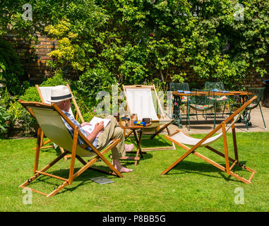 Ältere Mann, der Panama Hut Lösung Kreuzworträtsel im Liegestuhl sitzen im großen Garten in Sommerhitze, London, England, Großbritannien Stockfoto