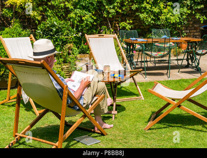 Ältere Mann, der Panama Hut Lösung Kreuzworträtsel im Liegestuhl sitzen im großen Garten in Sommerhitze, London, England, Großbritannien Stockfoto