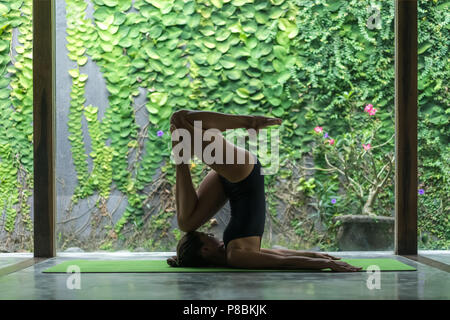 Seitenansicht des athletischen jungen Frau Yoga in Eingang in Pflug (Halasana) vor der Wand mit grünen Blättern bedeckt Stockfoto