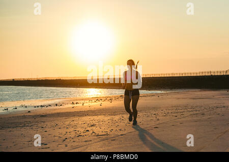 Silhouette der Sportlerin mit Smartphone im laufenden Armband Fall Jogging am Strand gegen Sonnenlicht Stockfoto