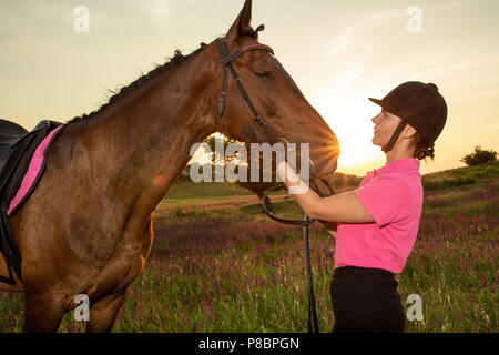 Schöne lächelnde Mädchen Jockey stand neben ihr braunes Pferd Tragen besonderer Uniform auf einen Himmel und grünen Feld Hintergrund auf einen Sonnenuntergang. Stockfoto