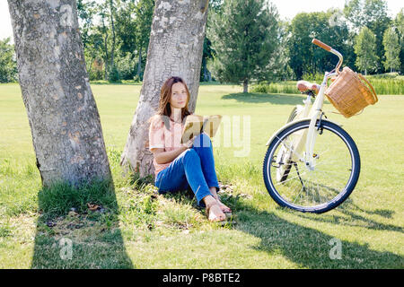 Schöne unbeschwerte Frau sitzt unter einem Baum mit einem Buch im Sommer Stockfoto