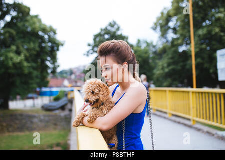 Porträt der Schönen lächelnden jungen Frau, die auf der Brücke mit ihrem kleinen roten Pudel Welpen. Stockfoto