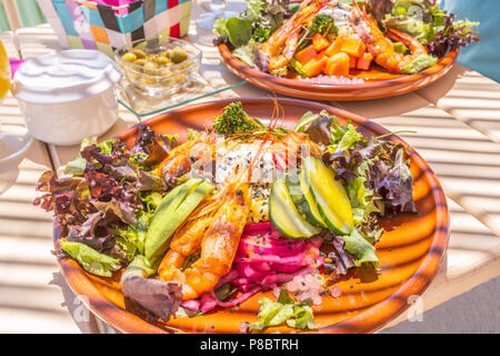 Zwei gemischte Salate auf einem weißen Tisch mit Streifen von einem Sonnenschirm in einer Beach Bar verursacht. Die gesunden Salat auf einer runden Platte hat Garnelen, Avocado, c Stockfoto