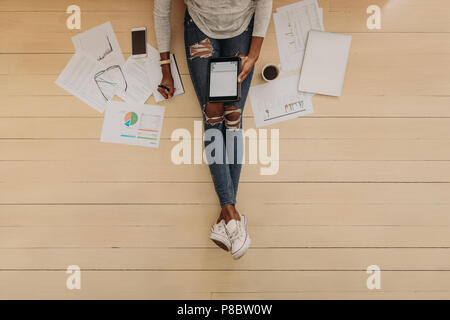 Blick von oben auf eine Frau im modischen Jeans arbeiten an Tablet PC zu Hause mit Kaffee und Geschäftspapiere an ihrer Seite sitzen. Frau Unternehmer sitzen Stockfoto