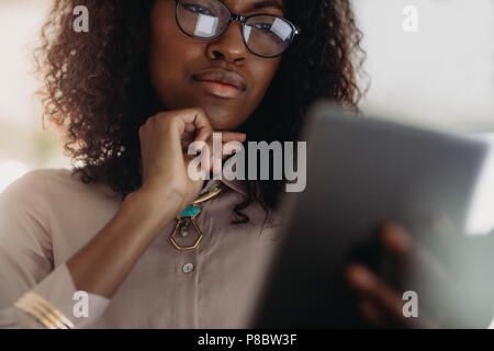 Nahaufnahme von einem lockigen Haaren Geschäftsfrau in Brillen bei Tablet-PC suchen mit der Hand am Kinn. Low Angle View der Unternehmerin in der formalen Kleidung l Stockfoto