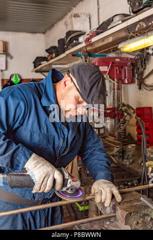 Ein junger Mann Schweißer in einem blauen T-Shirt, Schutzbrille und Handschuhe Prozesse Metall ein Winkelschleifer in der Garage, im Hintergrund eine Menge von Werkzeugen Stockfoto