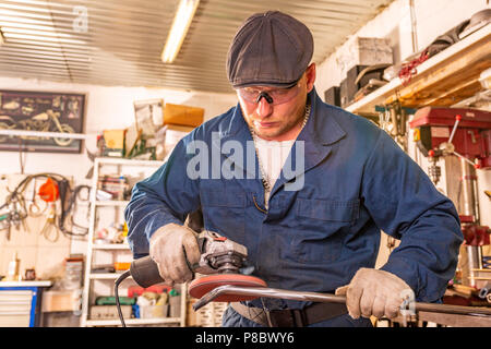 Ein junger Mann Schweißer in einem blauen T-Shirt, Schutzbrille und Handschuhe Prozesse Metall ein Winkelschleifer in der Garage, im Hintergrund eine Menge von Werkzeugen Stockfoto