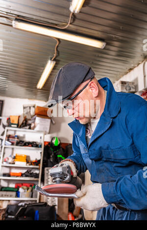 Ein junger Mann Schweißer in einem blauen T-Shirt, Schutzbrille und Handschuhe Prozesse Metall ein Winkelschleifer in der Garage, im Hintergrund eine Menge von Werkzeugen Stockfoto