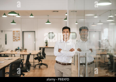 Lächelnd asiatische Geschäftsmann gegen eine Glaswand bei der Arbeit schiefen Stockfoto