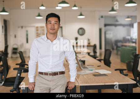 Zuversichtlich asiatische Geschäftsmann, lehnend auf einem Schreibtisch in einem Büro Stockfoto