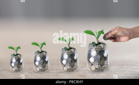 Hand eine Münze in ein Glas, die eine wachsende Pflanze in Münzen auf Holztisch mit unscharfen Hintergrund und kopieren Raum Stockfoto