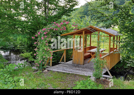 Pavillon aus Holz mit einem Dach und einem Tisch stehend auf die Bretter über den Bach unten liegen. Und wachsenden Blumen auf einem Bogen aus Draht gemacht in der Nähe. . Für yo Stockfoto