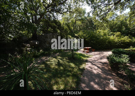 Kleine hölzerne Bänke mit Tisch im Schatten der Bäume in ein gemütliches Plätzchen im Park neben dem Pfad und einen großen Stein in der Nähe der Sträucher. Stockfoto