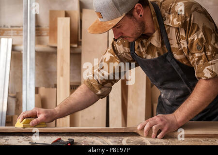 Ein Mann in einem Cap und Shirt poliert die Holzblock mit Sandpapier vor dem Lackieren in der Werkstatt, im Hintergrund, Werkzeuge und Bohren mach Stockfoto