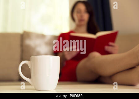 Frau mit Tasse heißen Tee lesen Buch von Fenster auf dem Sofa zu Hause. Fokus auf Tasse Tee Stockfoto