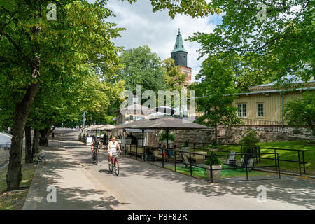 TURKU, FINNLAND - 8/7/2018: Leute auf einem Fahrrad neben den Fluss Aura im Sommer Tag mit dem Turm von Dom steigen über das Grün der Bäume. Stockfoto