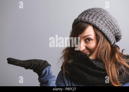 Verspielte attraktive junge Frau in einem warmen Woolly winter Outfit heraus halten Ihr leeres Palm mit einem glücklichen Lächeln für Product Placement und Präsentation Stockfoto