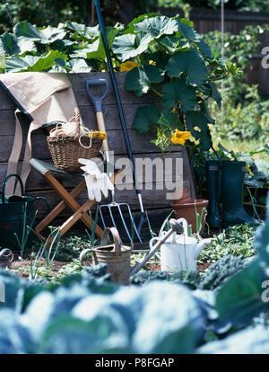 Garten Gabel neben einer Leinwand Hocker mit einem Vintage Warenkorb lehnte sich gegen einen rustikalen hölzernen Zaun in einem Gemüsegarten Stockfoto